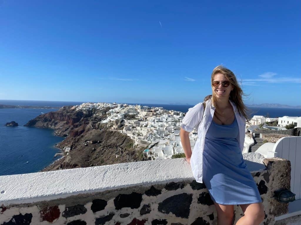 Woman sitting against ledge with white buildings and ocean in background 