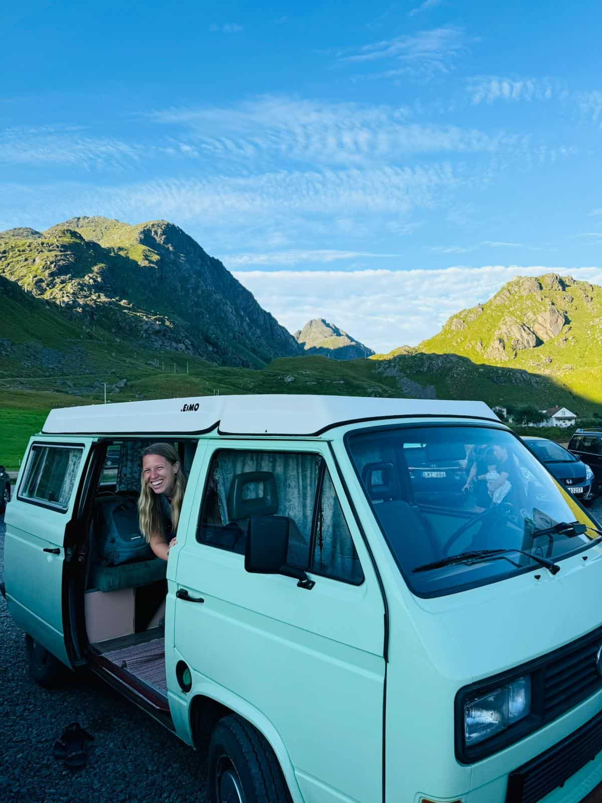 Woman sitting inside mint green Volkswagen van with mountains in background. 